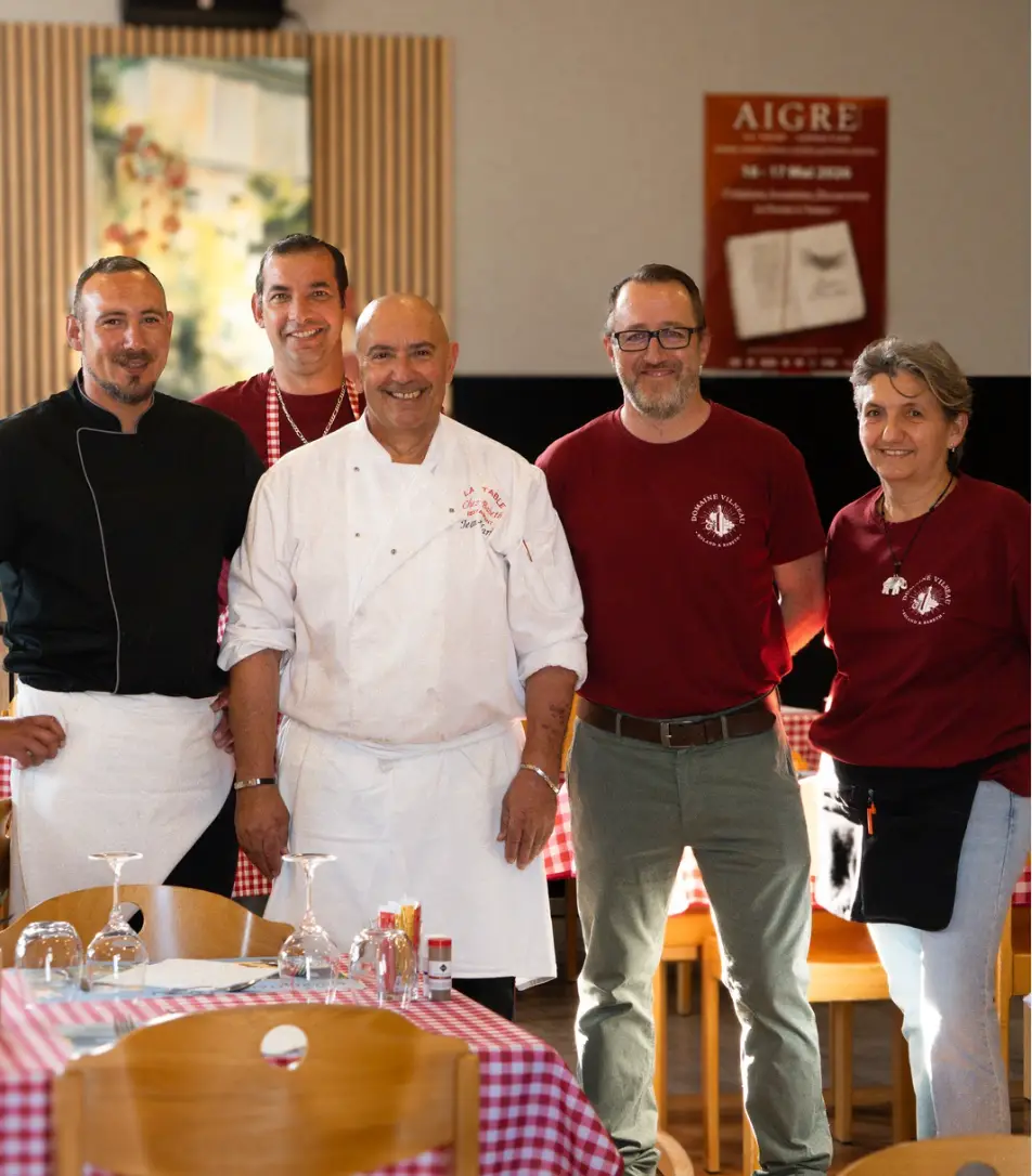 Photo d'équipe d'un restaurant, chef en veste blanche entouré du personnel en polo bordeaux devant un panneau « Aigre », portrait par Aptitude Pro.